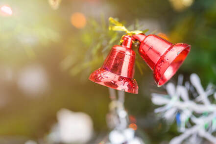 Close-up of two red Christmas bells hanging on a decorated Christmas tree, softly lit with warm golden holiday lights in the background to show the comforting sound of christmas songs played on holiday bells.