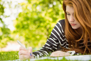 Red haired young woman lying on the grass on a warm fall day with a pen in hand studying with the sound of green noise around her to help her focus on her studying 