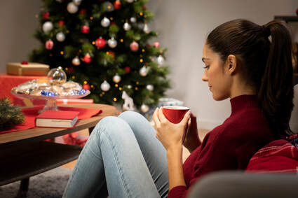 Young woman relaxing with a cup of tea in her living room decorated with holiday decorations and a christmas tree showing how calming Christmas music can help calm the nervous system and relieve holiday stress.
