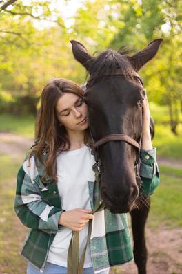 beautiful young woman cradles her horse's head to calm him while soothing ambient music plays in the background and dappled light sifts through the leaves of the green trees