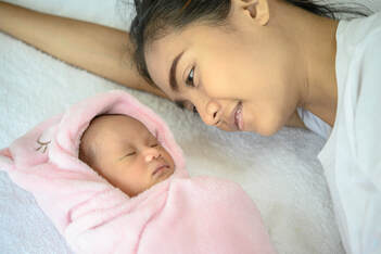 Mother lovingly watching her newborn baby sleep peacefully wrapped in a soft pink blanket -- calm sleep environment supported by pink noise.