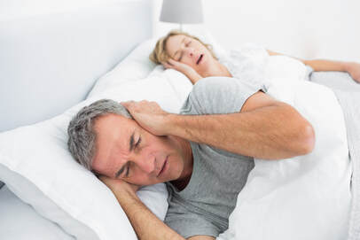Man holding his ears in bed while his wife lies next to him sleeping while snoring to show that white noise could help him sleep better