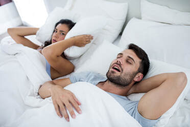 Young woman blocking her ears with her pillow while lyig next to her husband in bed who is snoring to show how white noise could help her sleep better