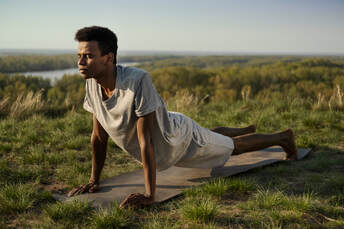 Young man practicing yoga outdoors in nature, holding upward-facing dog pose on a mat with a scenic forest and river view. Peaceful and grounding yoga practice in the fresh air.